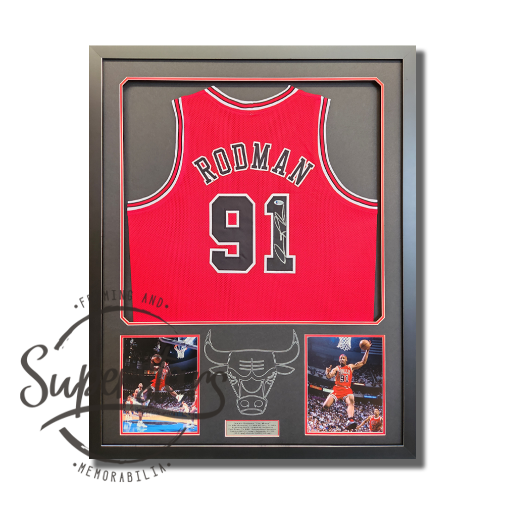 Dennis Rodman Chicago Bulls jersey signed by Rodman in silver ink across the black number 91. Below the jersey are two full colour action photos of Rodman playing for the Chicago Bulls. In between the photos is the outline of a bulls head and horns in silver pen. A silver plaque details his career in black writing. It has a black frame that has a silver metallic foil edges. The borders are black with a red trim.