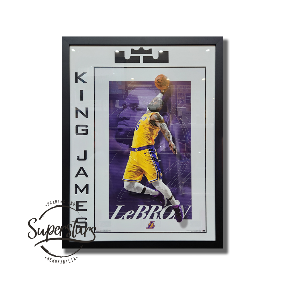 Lebron James Poster. Featuring Lebron's nick name 'King James' which has been cut into the matting and runs vertically down the left hand side. An icon of a crown has been cut into the matting across the top. This surrounds a full colour poster of Lebron about to slam dunk. The word LeBron is part of the print. This has a black trim line, white border and the icon and wording is black. Surrounded by black timber frame.