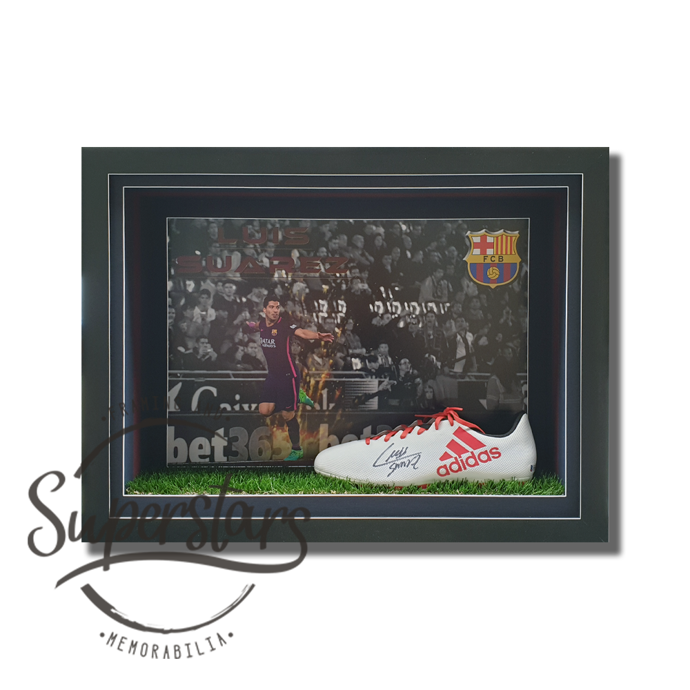 A soccer boot that has been signed sits on astro turf behind glass. In the background is an action photo of Luis Suarez playing. The frame is a shadow box with a black frame